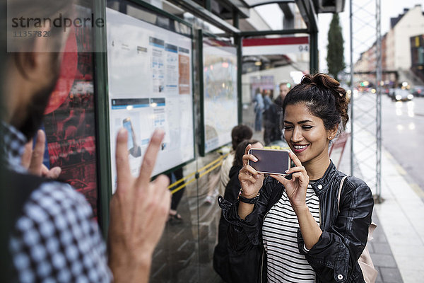 Lächelnde Frau fotografiert Mann durch Smartphone an der Bushaltestelle in der Stadt