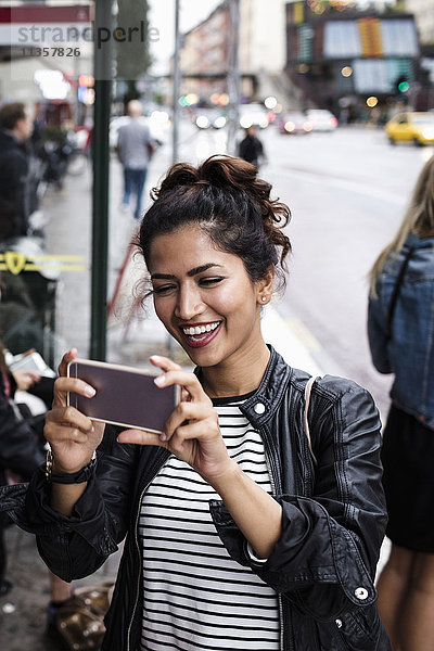 Glückliche Frau beim Fotografieren über Smartphone an der Bushaltestelle in der Stadt