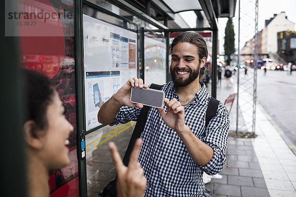 Lächelnder Mann fotografiert Freundin durch Smartphone an der Bushaltestelle in der Stadt