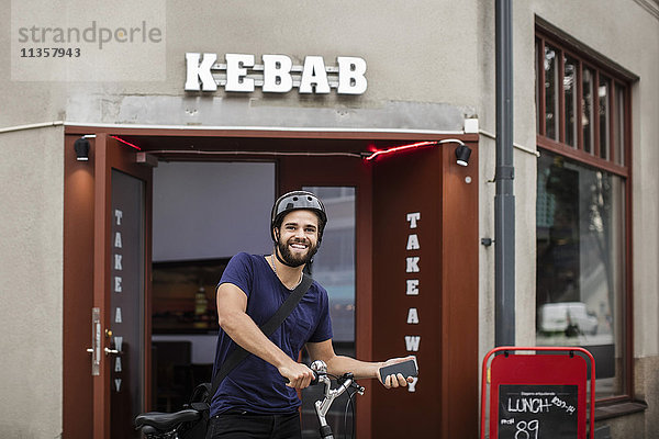 Porträt eines lächelnden Mannes mit einem Fahrrad  das ein Smartphone hält  während er gegen ein Restaurant in der Stadt steht.