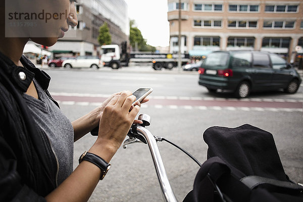 Abgeschnittenes Bild einer Frau mit Fahrrad auf dem Handy  während sie in der Stadt an der Straße steht.