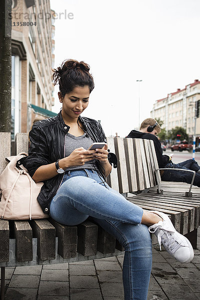 Lächelnde Frau beim Sitzen auf einer Holzbank in der Stadt mit dem Handy