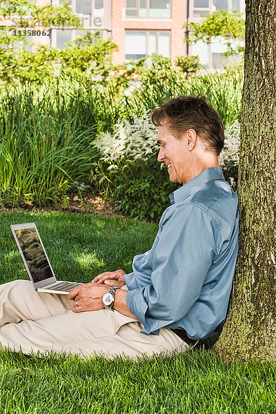 Geschäftsmann sitzt im Freien  an einen Baum gelehnt  mit Laptop