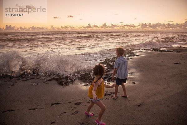 Junge und Schwester laufen bei Sonnenaufgang am Strand vor den plätschernden Wellen weg  Blowing Rocks Preserve  Jupiter Island  Florida  USA