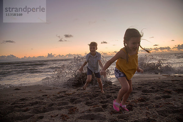 Junge und Schwester laufen bei Sonnenaufgang vor den plätschernden Wellen weg  Blowing Rocks Preserve  Jupiter Island  Florida  USA