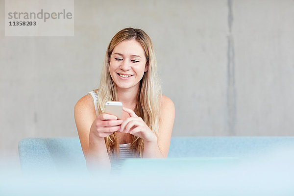 Junge Studentin sitzt auf einem Sofa im Studienraum und benutzt ein Smartphone an einer Hochschule