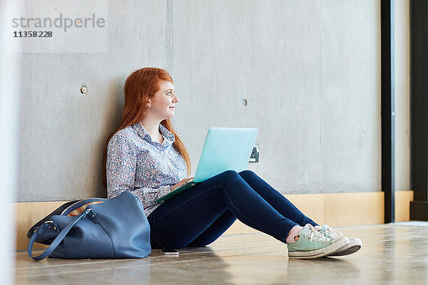 Junge Studentin mit Laptop auf dem Boden sitzend an einer Hochschule