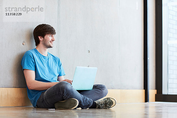Junger männlicher Student mit Laptop auf dem Boden sitzend an einer Hochschule