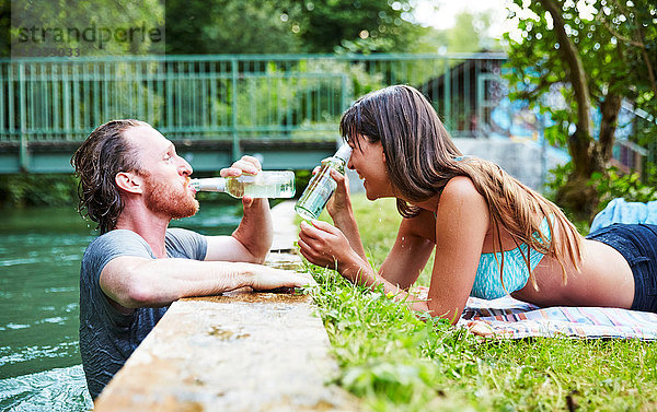 Junger Mann im Fluss  junge Frau liegt im Gras am Flussufer  Mann trinkt aus Bierflasche