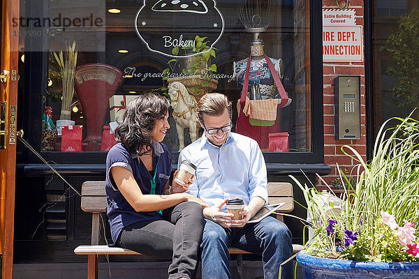 Paar auf Bank sitzend  vor Bäckerei  Kaffeetassen haltend