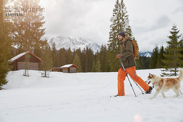 Mittelgroßer erwachsener Mann auf Schneeschuhen durch verschneite Landschaft  Hund neben ihm  Elmau  Bayern  Deutschland