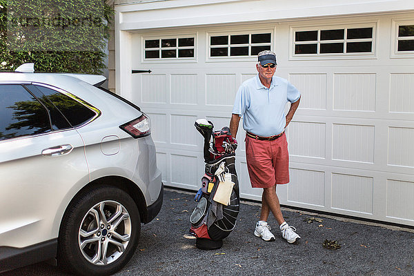 Porträt eines älteren männlichen Golfspielers  der mit einer Golftasche neben dem Garagentor steht