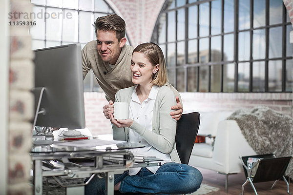Junge Frau zu Hause  am Schreibtisch sitzend  auf den Computerbildschirm schauend  Mann neben ihr  lächelnd