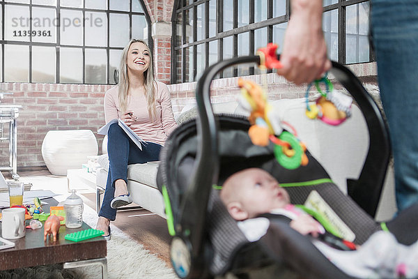 Vater verlässt das Haus mit Mädchen im Autositz  Mutter sitzt auf dem Sofa und arbeitet