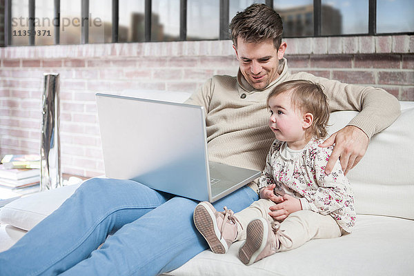 Vater sitzt mit seiner kleinen Tochter auf dem Sofa und schaut auf den Laptop