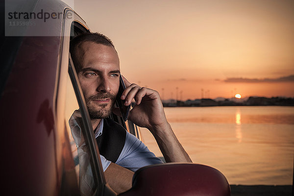 Geschäftsmann am Autofenster im Gespräch mit Smartphone bei Sonnenuntergang an der Küste  Cagliari  Sardinien  Italien