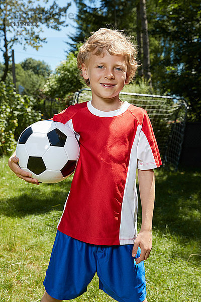 Porträt eines Jungen in Fussballuniform  der im Garten einen Fussball hält