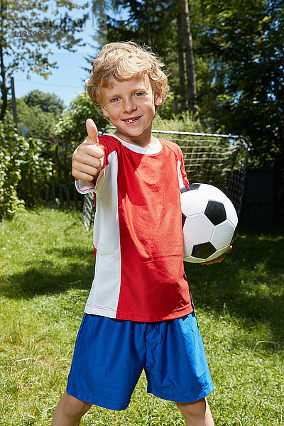 Porträt eines Jungen in Fussballuniform  der im Garten einen Fussball hält und Daumen hochhebt