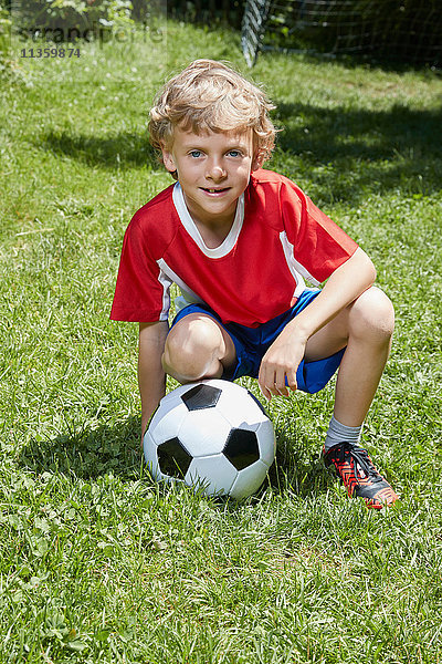 Porträt eines Jungen in Fussballuniform  der mit Fussball im Garten kauert