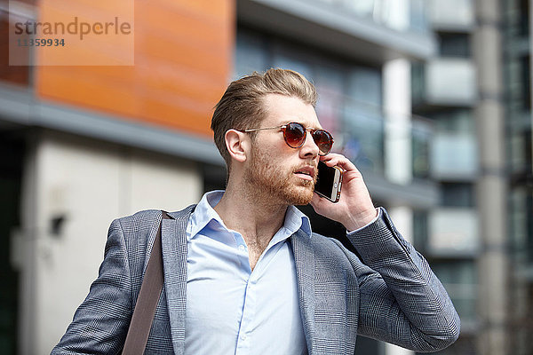 Junger Geschäftsmann mit Sonnenbrille spricht auf dem Smartphone vor dem Büro  London  UK