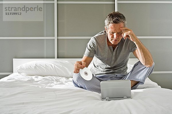 Älterer Mann sitzend  CD haltend und auf den Laptop starrend