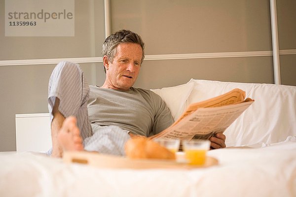Älterer Mann sitzt im Bett und liest Zeitung.