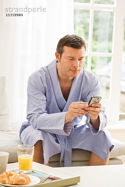Der reife Mann auf dem Sofa liest Texte auf dem Handy und frühstückt.