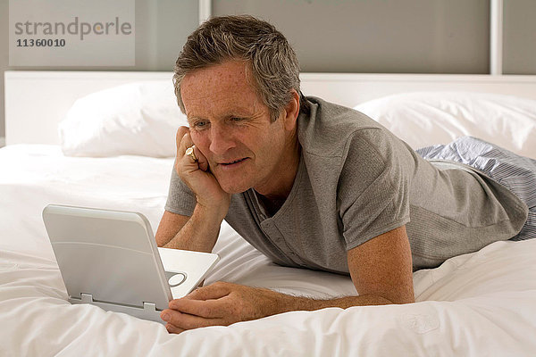 Verwirrter älterer Mann  der auf dem Bett liegt und auf den Laptop starrt.
