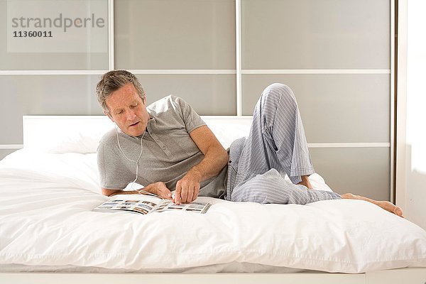 Senior Mann liegt auf dem Bett  liest Magazin und hört Kopfhörer.