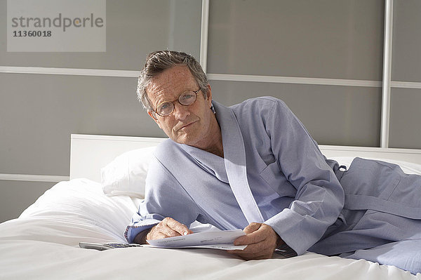 Porträt eines älteren Mannes  der auf dem Bett liegt und Rechnungen prüft.