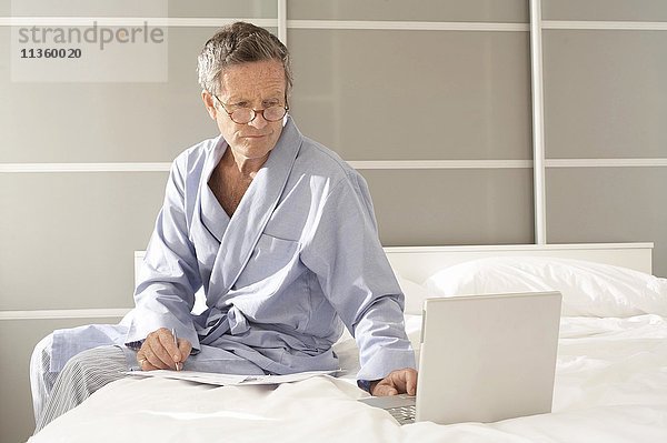 Senior Mann sitzt auf dem Bett und prüft Rechnungen auf dem Laptop.