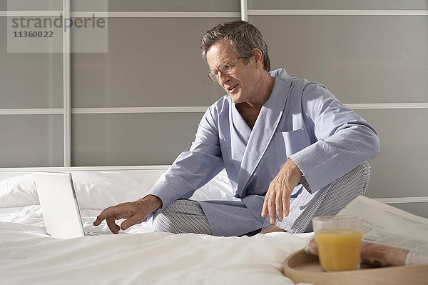 Verwirrter älterer Mann auf dem Bett  der auf dem Laptop schreibt.
