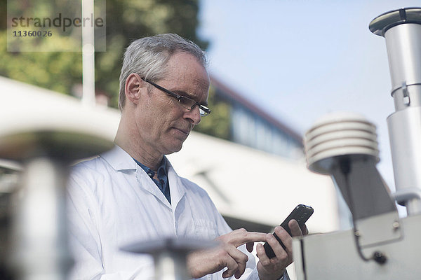 Männlicher Meteorologe schreibt auf Smartphone an Wetterstationsausrüstung im Freien