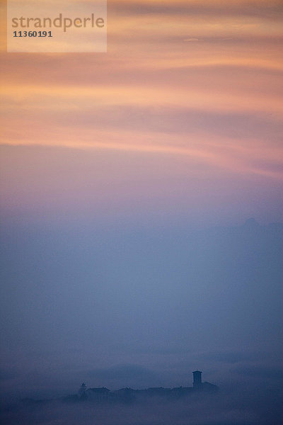 Silhouettierte Dächer  die bei Sonnenuntergang aus niedrigen Wolken auftauchen  Langhe  Piemont. Italien