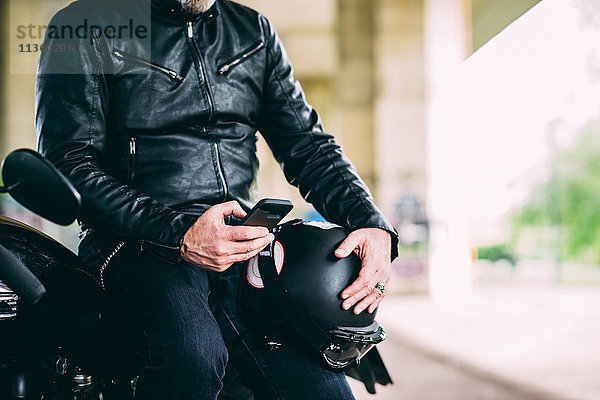 Mittelteil eines reifen Motorradfahrers  der auf dem Motorrad sitzt und auf dem Smartphone texten kann.