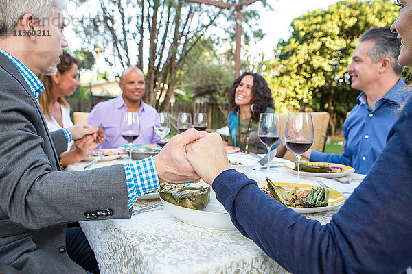 Sechs reife erwachsene Freunde halten sich am Gartenparty-Tisch an den Händen