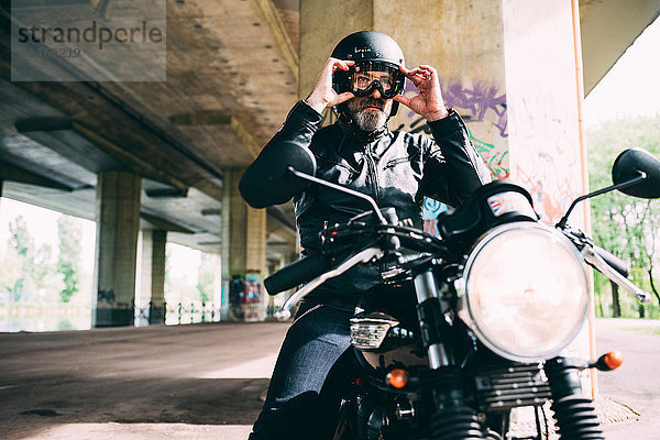 Erwachsener Motorradfahrer  der auf dem Motorrad sitzt und unter der Überführung eine Schutzbrille aufsetzt.