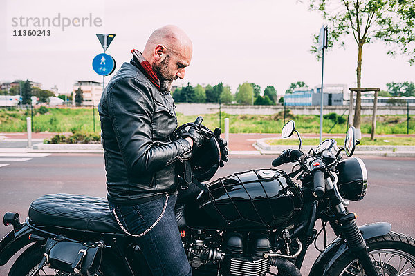 Erwachsener Motorradfahrer  der auf dem Motorrad sitzt und den Helm untersucht.