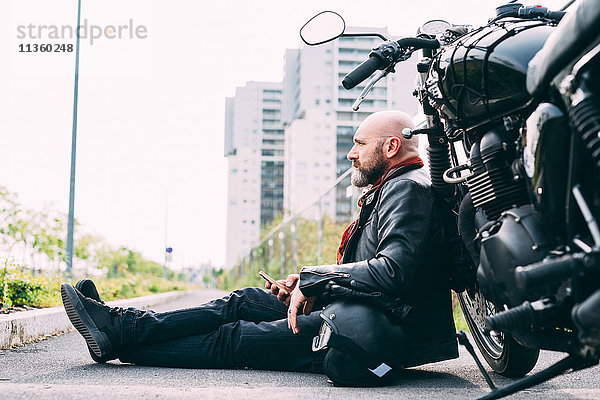 Erwachsener Motorradfahrer am Straßenrand sitzend  auf dem Motorrad lehnend  mit Smartphone