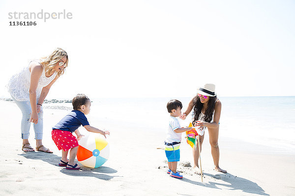 Zwei Mütter und kleine Söhne spielen mit Beachball und Windrädern am Strand  Malibu  Kalifornien  USA