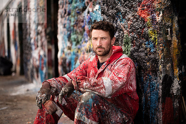 Porträt eines männlichen Schiffsmalers  der gegen eine mit Farbe bespritzte Wand sitzt