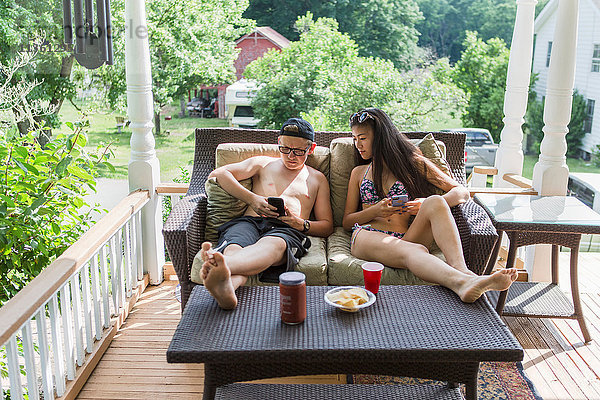 Teenager-Paar im Bikini und in Badeshorts auf dem Terrassensofa liegend  lesen gemeinsam auf einem Smartphone