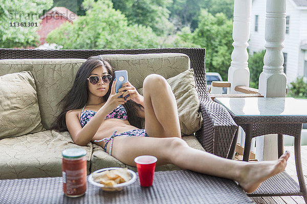 Teenager-Mädchen im Bikini auf Terrassensofa liegend und Smartphone lesend