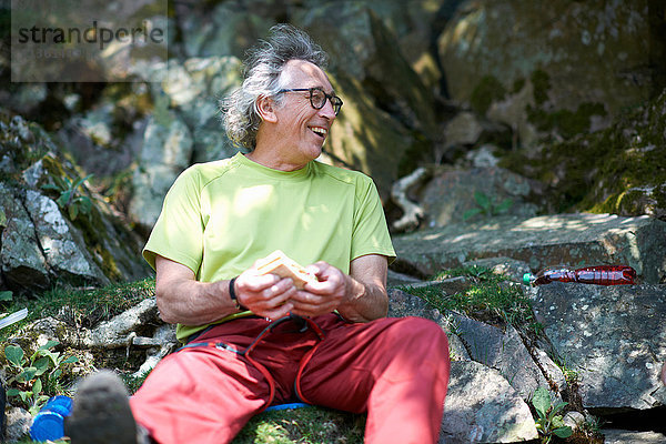 Wanderer sitzt auf Felsen und hält ein Sandwich in der Hand und schaut lächelnd weg