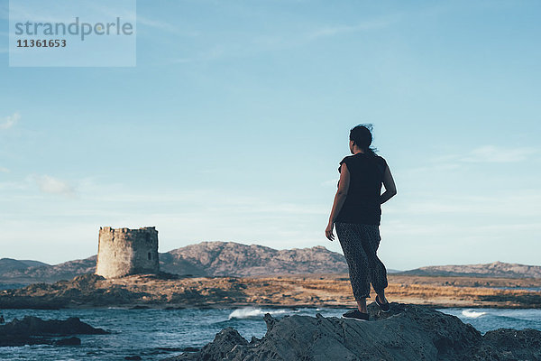 Rückansicht einer Frau auf Felsen mit Blick über das Meer auf einen verfallenen Leuchtturm  Stintino  Sassari  Italien