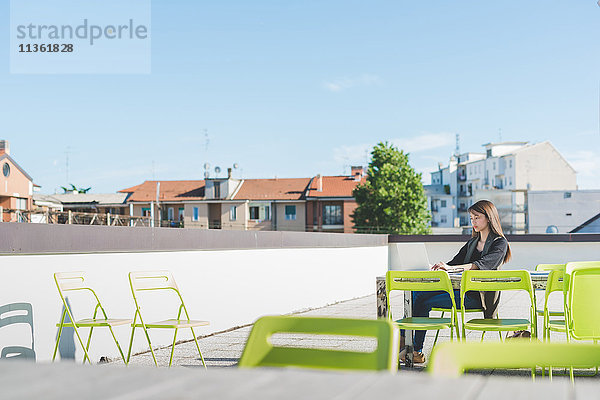 Junge Frau sitzt auf der Dachterrasse der Stadt und tippt auf dem Laptop.