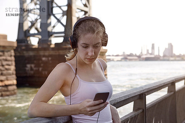 Junge Frau am Fluss  Kopfhörer tragend  mit Smartphone  New York City  USA