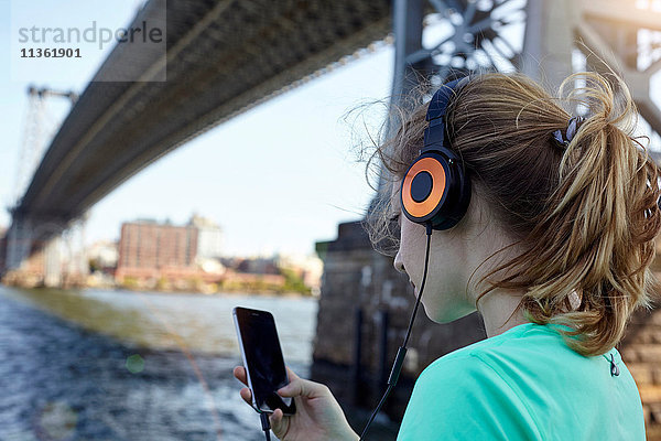 Junge Frau am Fluss  Kopfhörer tragend  Smartphone benutzend  Rückansicht  New York City  USA