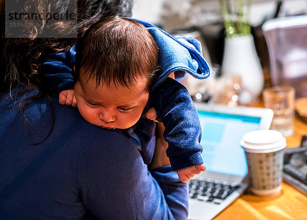 Mutter wiegt den neugeborenen Sohn und benutzt den Laptop an der Küchenbar.