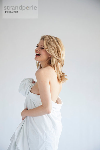 Studioaufnahme einer schönen  nackten  blonden  jungen Frau in ein Laken gehüllt  lachend